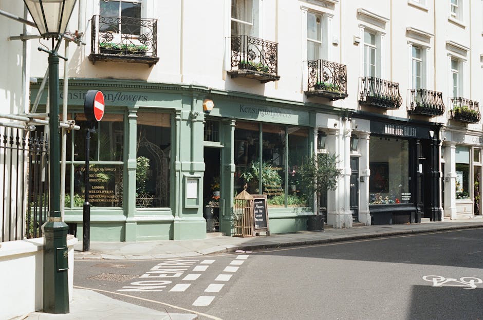 Photograph of a street-facing retail store with a soft green facade and large glass windows showcasing floral arrangements and decorative plants inside. The building features several small balconies w