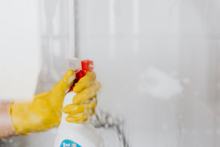 Close-up image of a person wearing yellow rubber gloves holding a white spray bottle while cleaning a glass surface, likely a household window or mirror. The surface appears clear and spotless, reflec