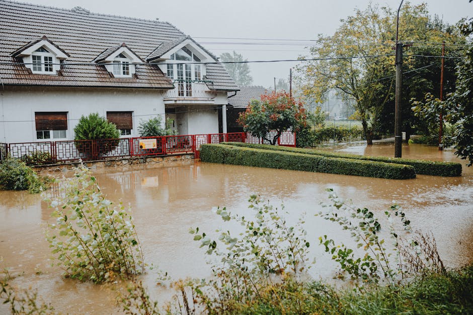 A residential front yard flooded with muddy water from heavy rain, partially submerging the grass and flower beds. The house in the background has white walls, multiple windows, and a tiled roof, with