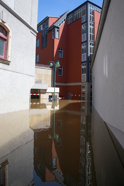 A residential front yard flooded with muddy water from heavy rain, partially submerging the grass and flower beds. The house in the background has white walls, multiple windows, and a tiled roof, with